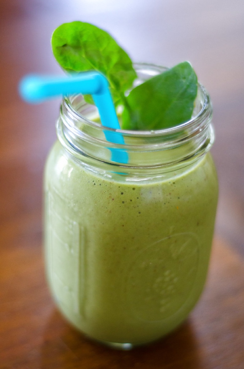 A glass of green smoothie with a swirl of peanut butter, next to a frozen banana and a handful of spinach