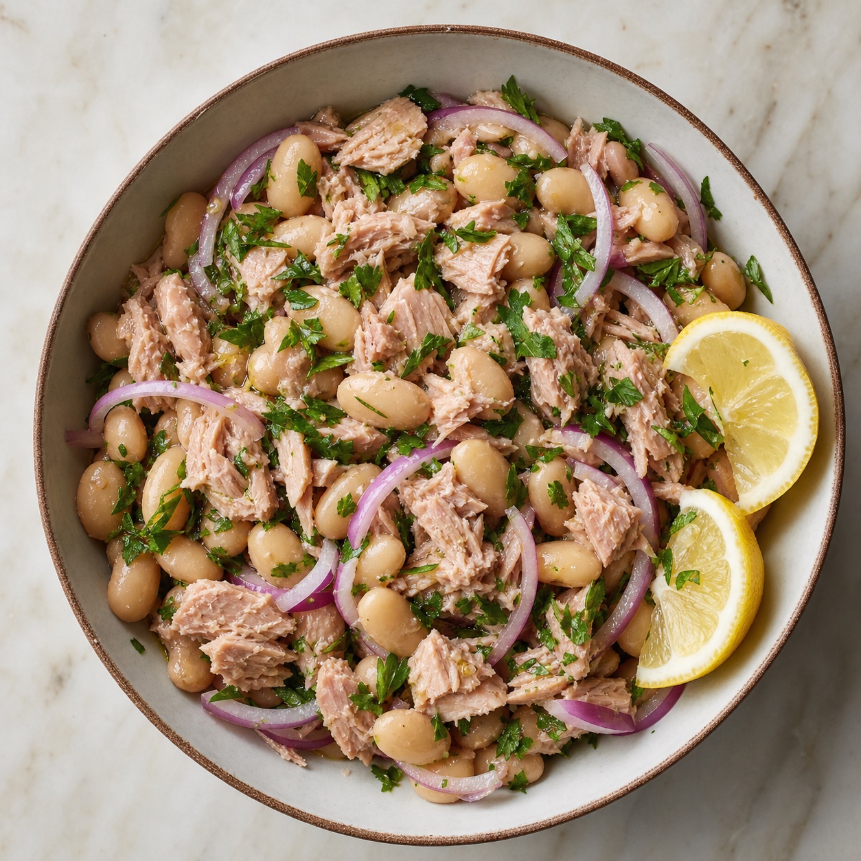 Overhead view of a tuna and white bean salad with red onion, parsley, and lemon wedges in a shallow bowl